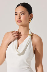 A woman is wearing a sleeveless white top with a draped neckline. She has gold earrings, a gold ring, and a gold bracelet. The accessories have a shiny, metallic finish.